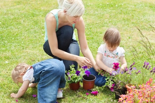 A woman with short blonde hair wearing a light blue sleeveless top and dark jeans is kneeling on a well-maintained grassy lawn in a garden, tending to potted plants and flowers. She is using her hands to adjust or plant the greenery in the soil. To her left, a young child with light brown hair, dressed in a white top and pink shorts, is sitting on the grass, holding a flower and observing the garden. Another child with curly blonde hair, wearing a blue dungaree, is leaning forward on the grass, exploring or picking flowers nearby. The garden features lush, neatly trimmed grass, with potted plants and flower beds filled with vibrant pink and purple blossom plants. In the background, there are garden borders and possibly a hedge, indicating a landscaped outdoor space. The scene suggests a peaceful, sunny day suitable for outdoor gardening and family activities, aligning with services such as lawn mowing and garden maintenance offered by Lawn Mowing Norwood in the Norwood area, perhaps within the local postcode region.